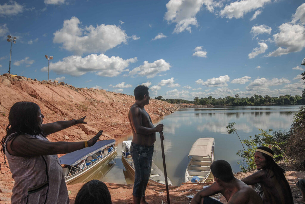 Uma mulher indígena estende os braços em direção ao rio. Ao lado, há um banco de terra vermelha. Outros indígenas estão ao redor da mulher.