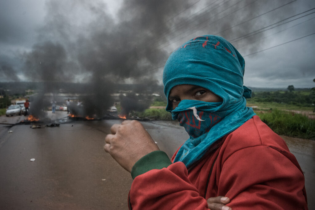Um manifestante usa uma camiseta azul como máscara, tampando todo seu rosto. Atrás dele, pneus queimam.