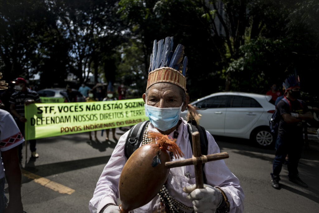 Indígena segura uma cruz e um instrumento, usando um cocar e uma máscara. Ao fundo, lê-se: STF defendam nossos direitos