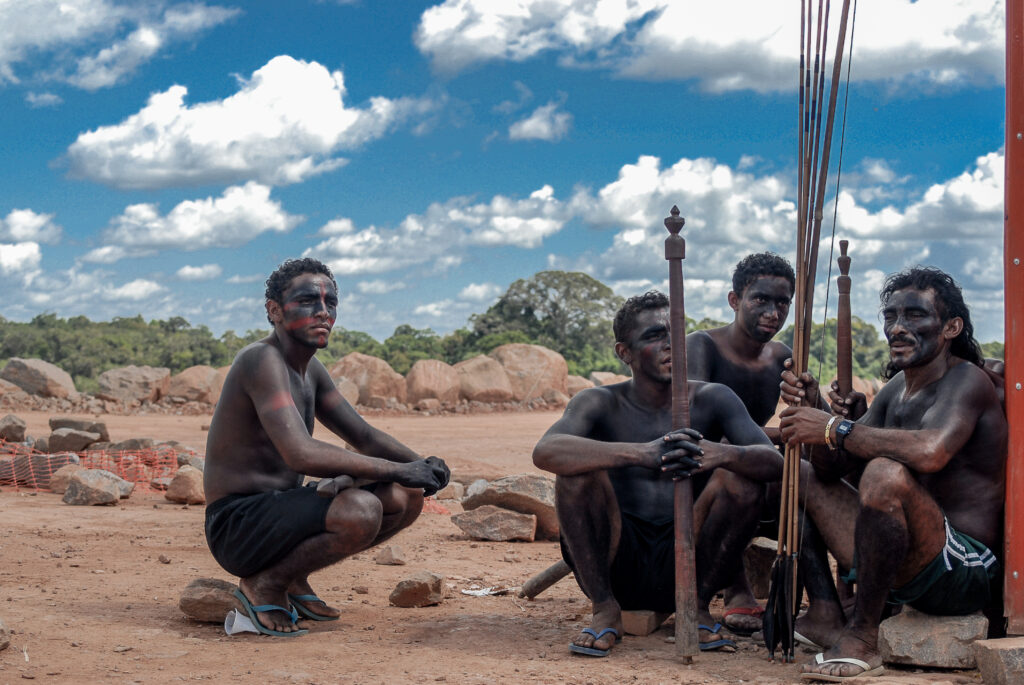 Grupo de indígenas pintados de tinta preta e vermelha sentados segurando flechas.