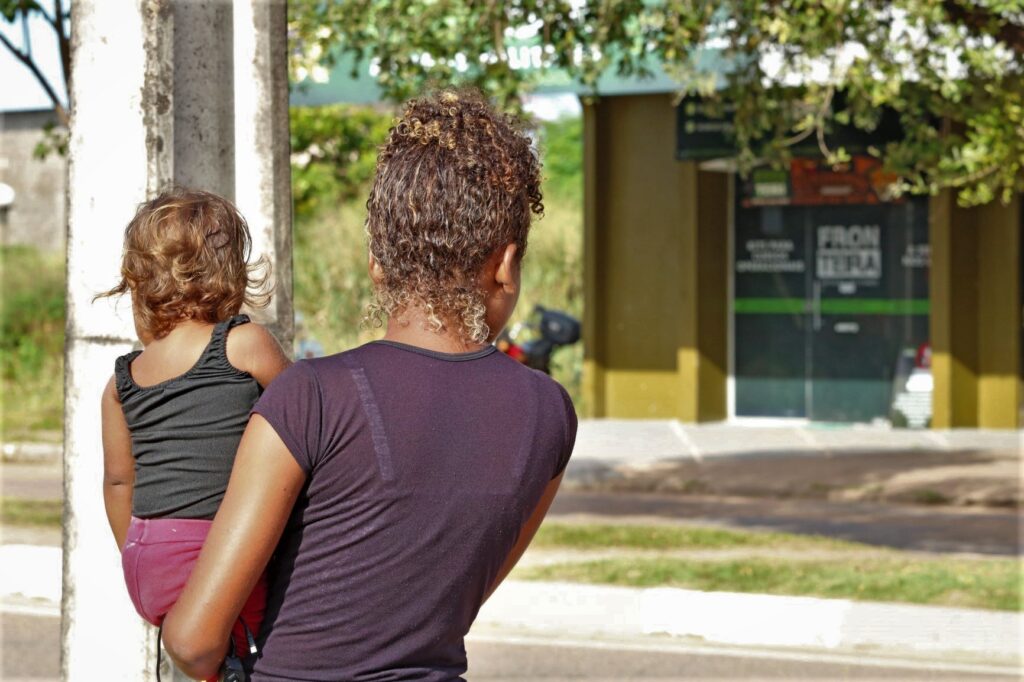 Uma mulher negra segura sua filha no colo. Ambas estão de costas
