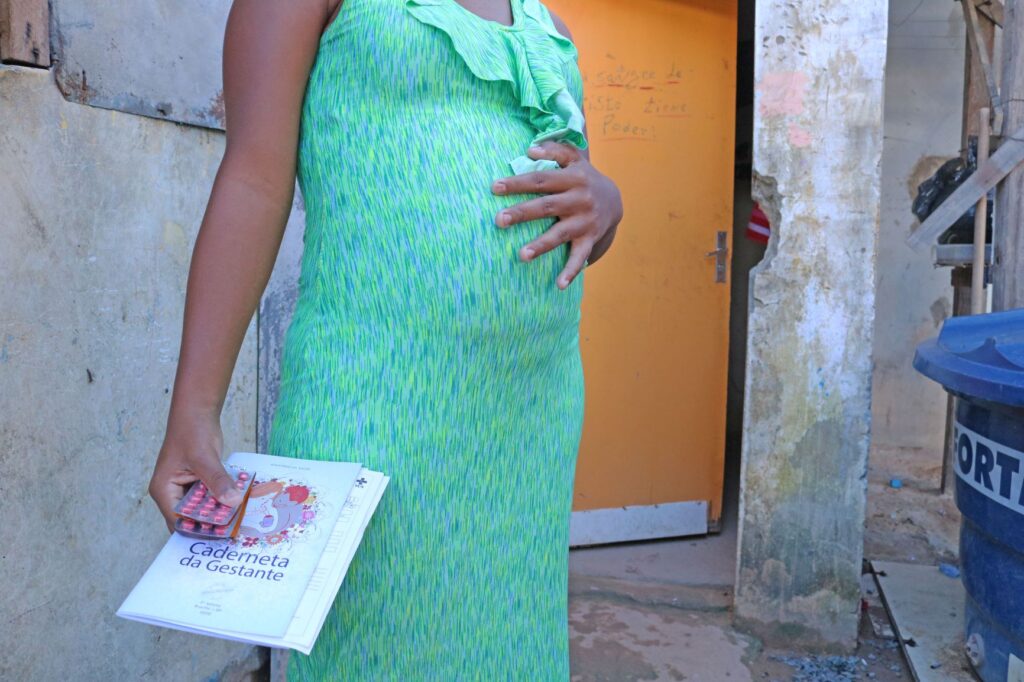 Uma grávida de vestido verde com a mão na barriga. Ela segura uma caderno escrito Caderneta da Maternidade