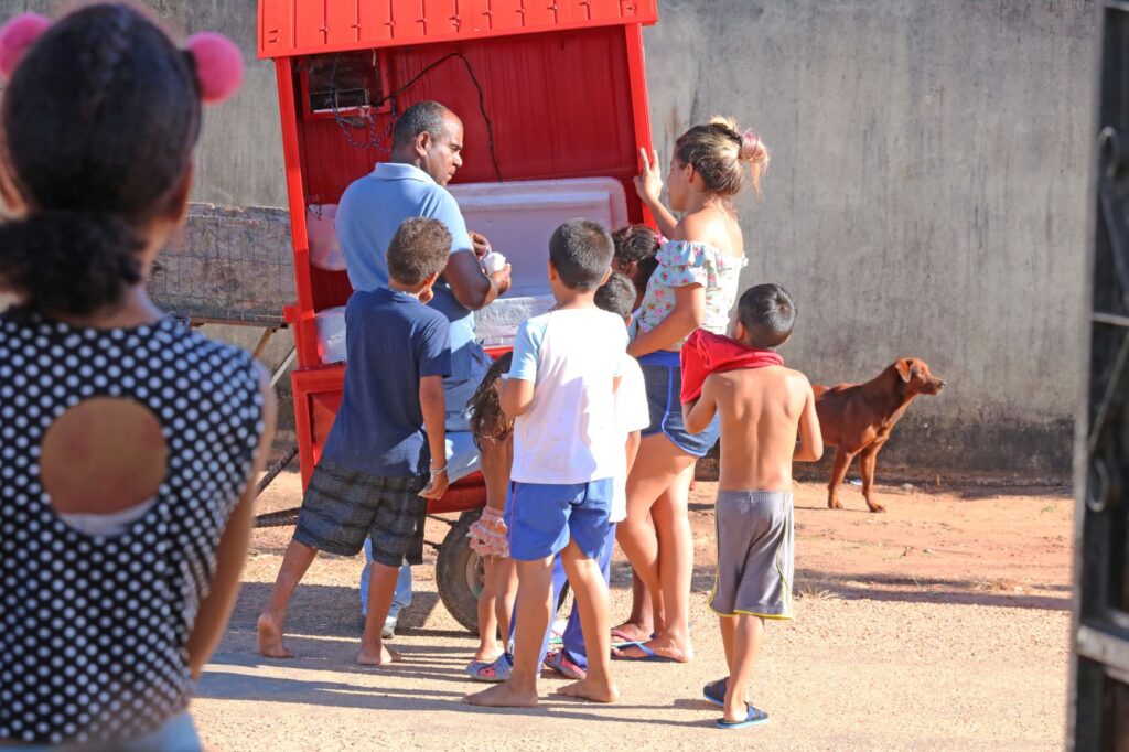 Crianças se amontoam junto de um vendedor de sorvete em um carrinho vermelho.