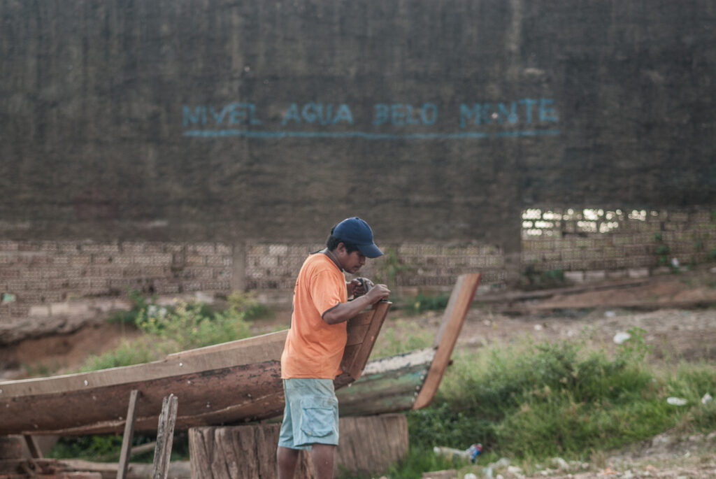 Um homem ao lado de uma canoa na terra firme. Ao fundo, está escrito "Nível Água Belo Monte"