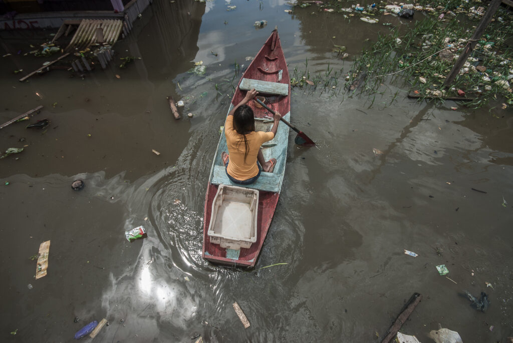 Uma mulher rema em um barco. No rio, há lixo ao redor