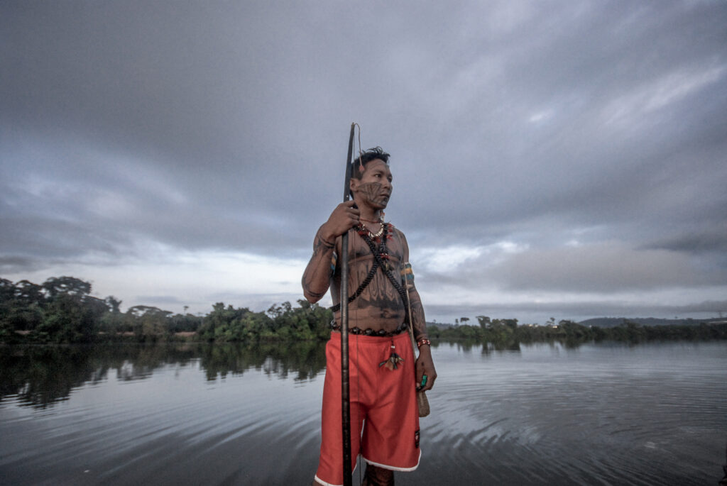 Um indígena de pé em uma canoa no meio do rio. Ele está sem camiseta e segura um arco grande.