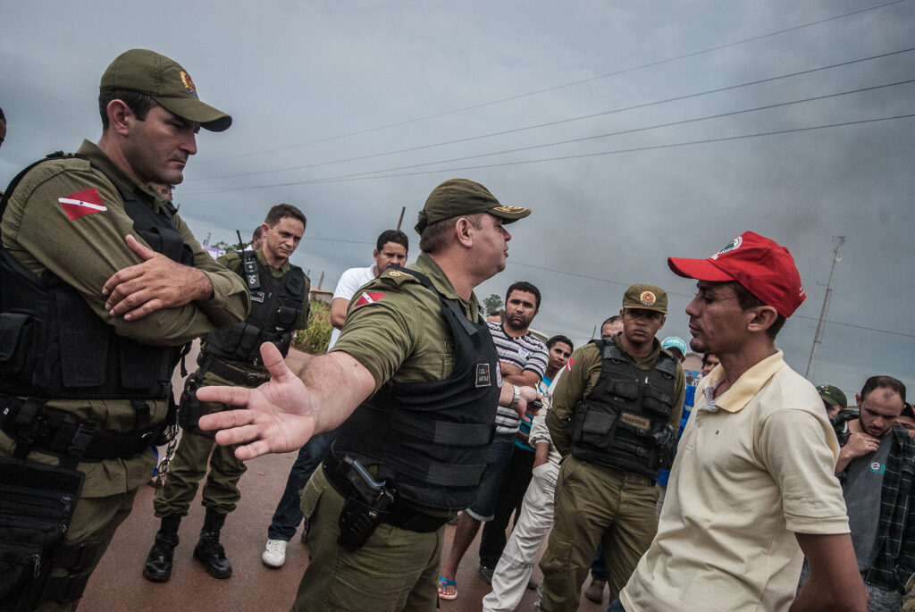 Policial fardado do lado esquerdo abre os braços enquanto questiona um manifestante