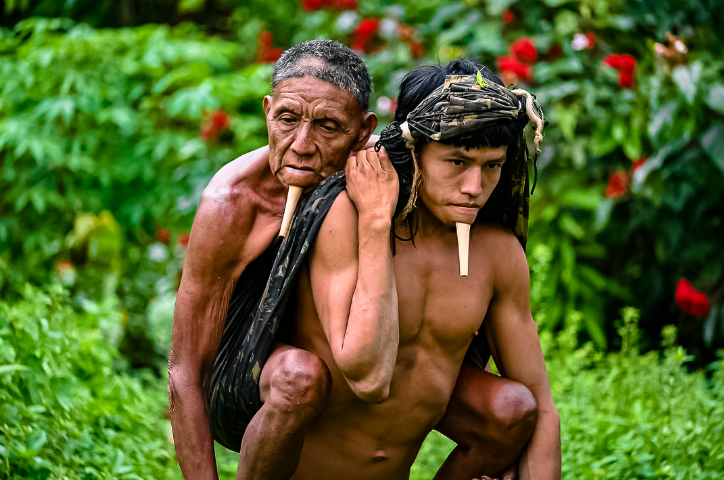imagem de um indígena jovem carrega um idoloso nas suas costas. Ao fundo, há vegetação