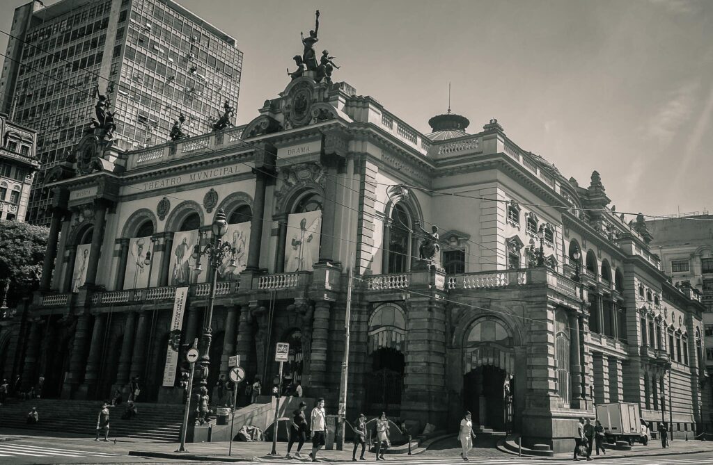 Foto em preto e branco do Teatro Municipal de São Paulo. Prédio antigo., com várias pessoas na rua.