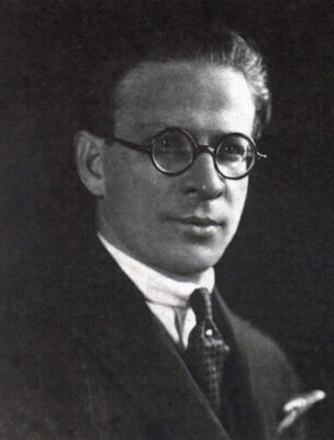 O retrato em preto e branco mostra o escritor Menotti Del Picchia, um homem branco de cabelos lisos e curtos penteados para trás, que usa óculos de grau de armação preta e redonda. Ele veste terno e gravata e posa de perfil, virado para a esquerda.