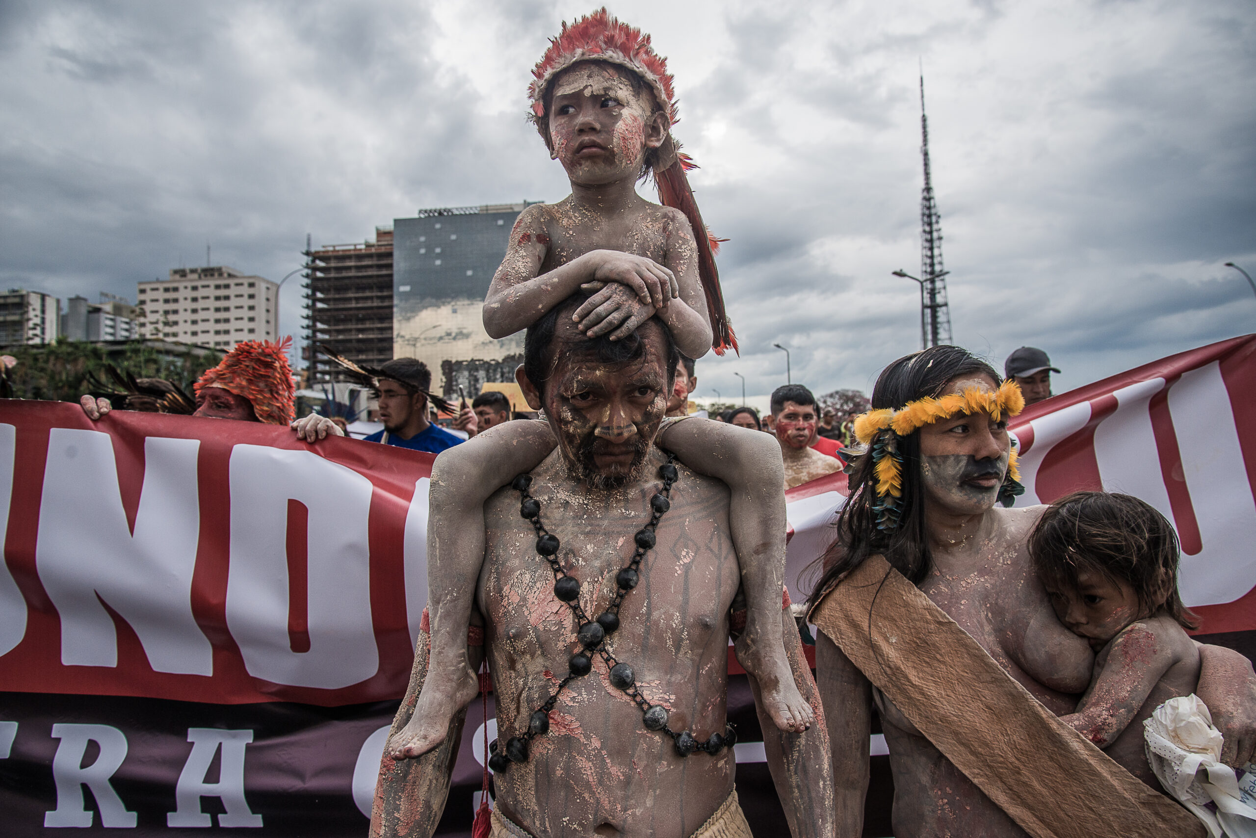 Criança indígena sobre os ombros de um homem adulto em protesto em Brasília. Ambos estão pintados de terra e urucum. Do lado direito, um bebê mama no peito da mãe.