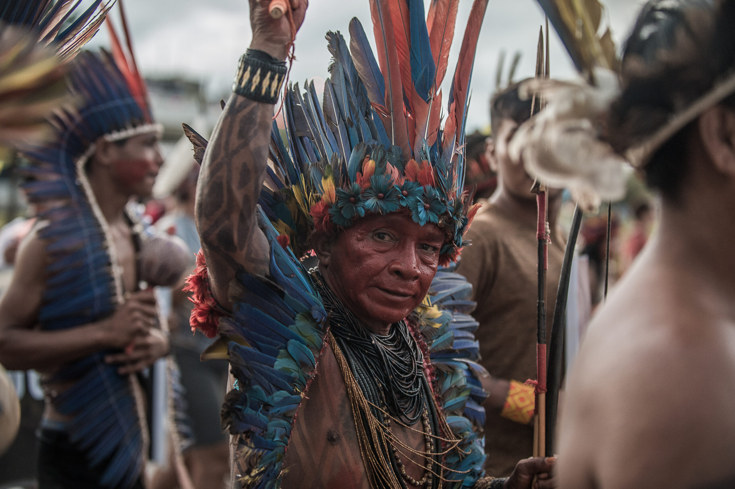 Homem indígena mais velho com cocar azul e vermelho ergue o punho ao alto no Acampamento Terra Livre