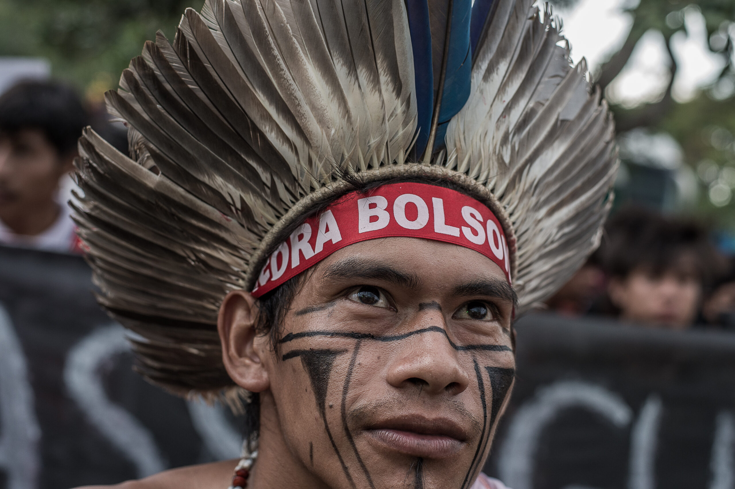 Homem indígena usando um cocar marrom, preto e branco olha ao longe. Ele tem pinturas geométricas pretas no rosto e usa uma faixa na testa vermelha, onde lê-se "Fora Bolsonaro".