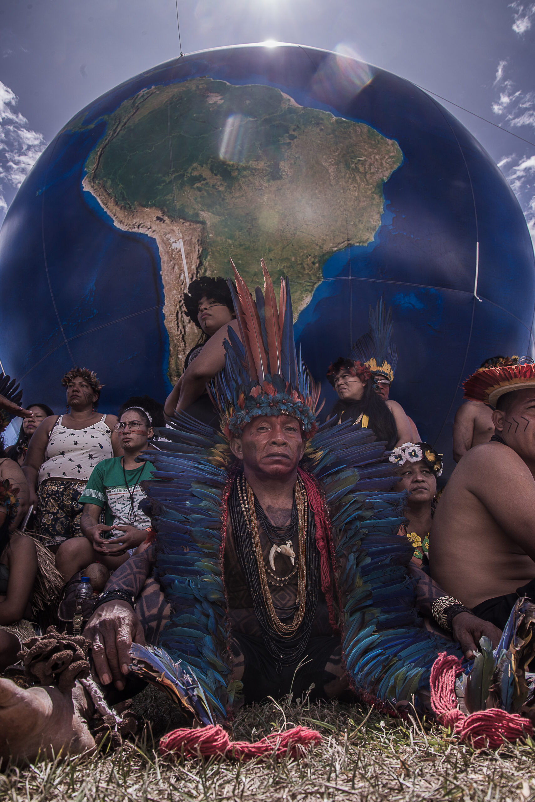 Grupo de manifestantes indígenas do Acampamento Terra Livre em volta de um globo terrestre inflável grande. No centro, um homem mais velho usando um cocar azul até os pés olha para a câmera com distinção. Homens, mulheres e crianças se aglutinam ao seu redor.