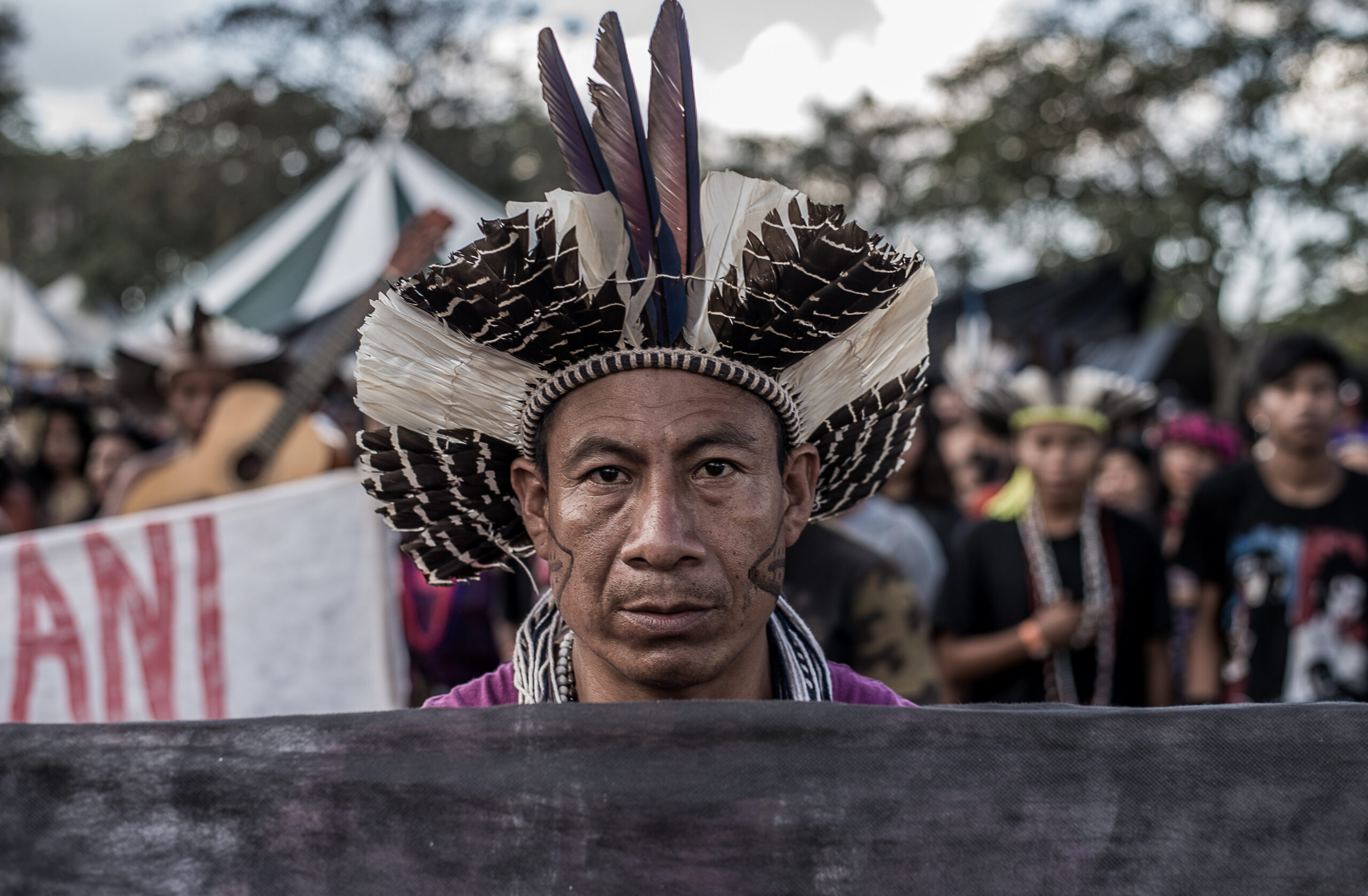 Homem indígena usa cocar marrom e branco e olha para a câmera atrás de um cartaz no protesto do Acampamento Terra Livre.