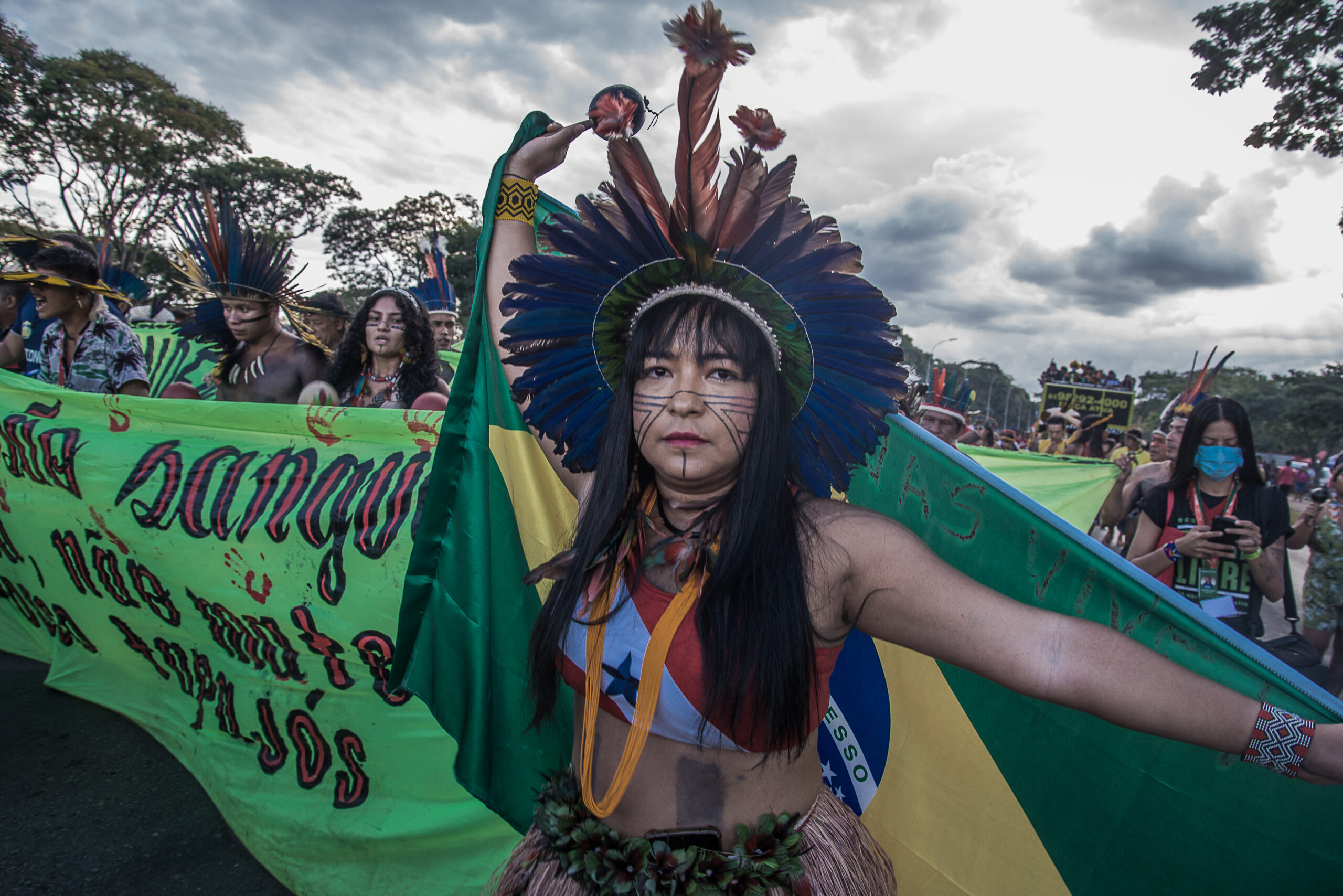 Mulher indígena usando um cocar marcha e ergue uma bandeira do Brasil nas costas no protesto do Acampamento Terra Livre.