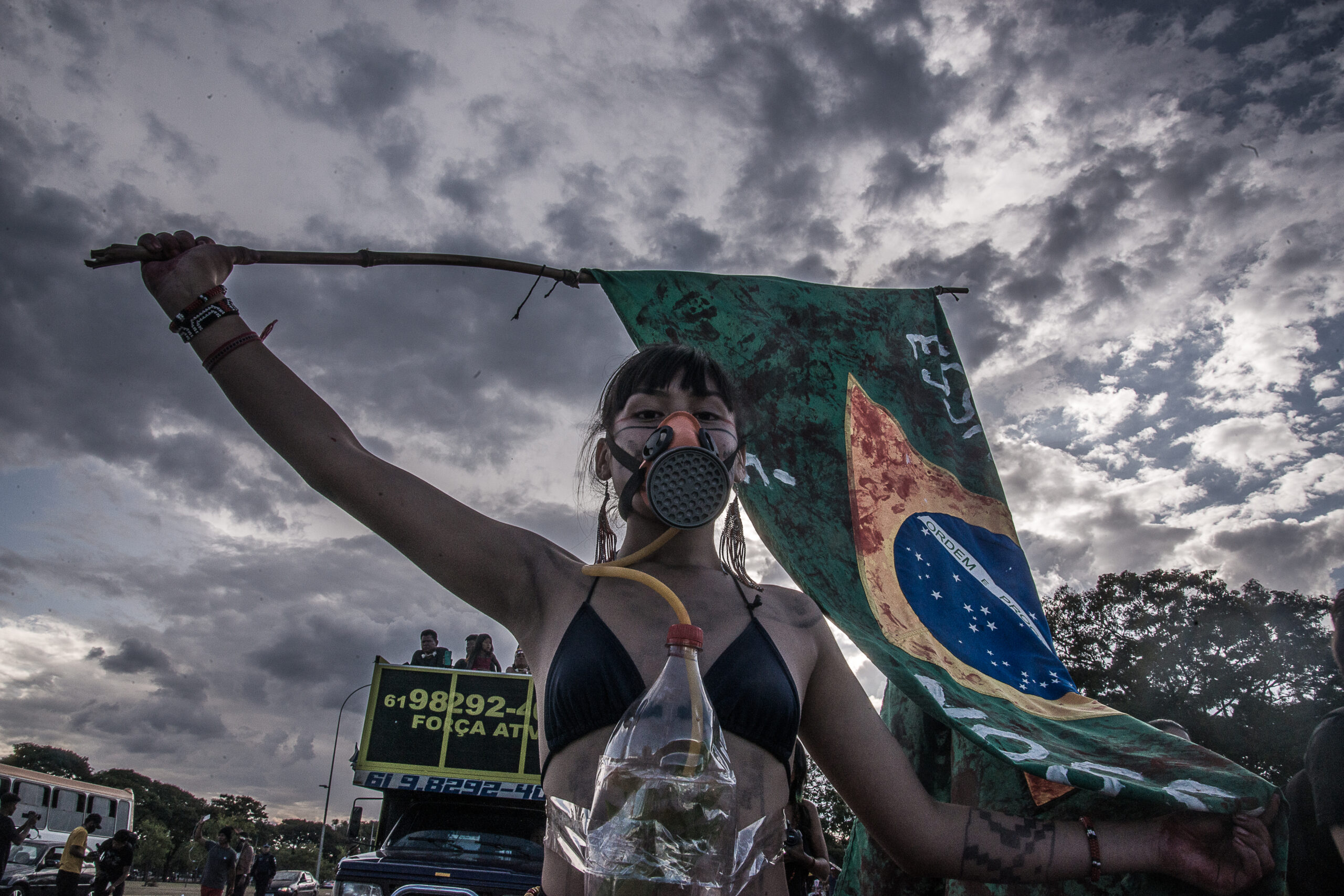 Mulher indígena segura uma bandeira do Brasil pintada de vermelho pela haste. Ela usa uma representação de máscara de gás, feita de garrafa pet.