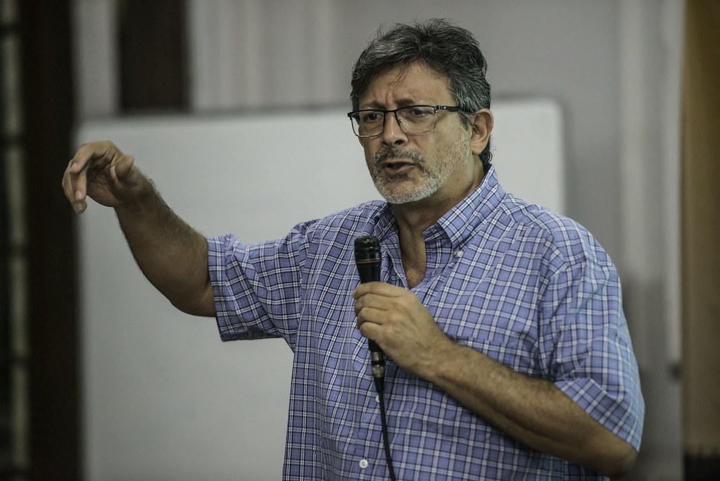 O professor Carlos Eduardo Young, da UFRJ, fala em um microfone em palestra. Ele usa uma camisa azul de mangas curtas.