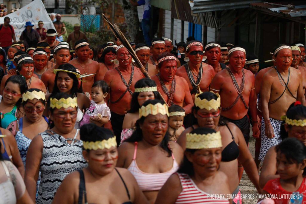 Mulheres indígenas vestem uma coroa de tecido; atrás homens indígenas pintados de vermelho, em pé