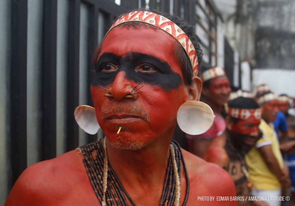 um indígena com olhos pintados de preto e rosto vermelho, olha sério para frente.