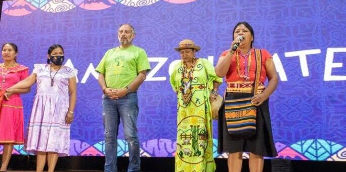 Palestrantes falam na 2ª Assembleia Mundial pela Amazônia. O palco é dominado por quatro mulheres, uma delas segurando o microfone.