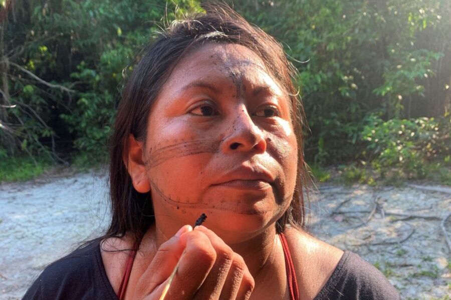 Na foto, Alessandra Korap, liderança Munduruku do Médio Tapajós, no Pará, recebe pintura facial. Ela fixa o olhar em algo distante, virando os olhos para a direita, enquanto uma mão com pincel faz traços com tinta preta em seu queixo