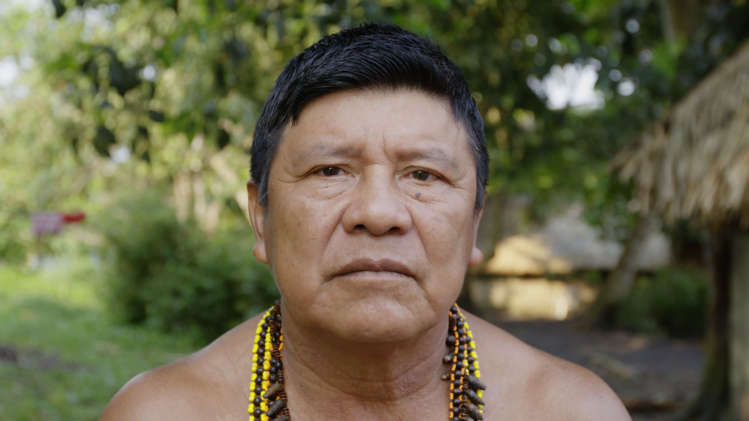 O cacique Jairo Saw, do povo Munduruku, olha diretamente para a câmera. O close mostra seu rosto, o peito nu e os colares coloridos em seu pescoço.