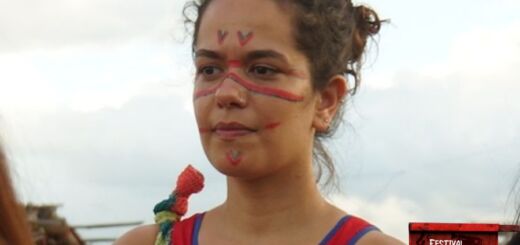 Close da antropóloga Ana Estrela semi-perfilada, olhando para a esquerda. Ela é uma mulher branca de cabelos e olhos castanhos. Ela está de coque e usa uma regata vermelha, e tem o rosto pintado com linhas vermelhas e azuis.