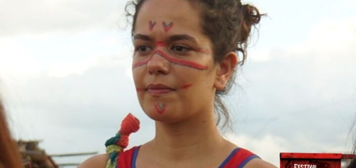 Close da antropóloga Ana Estrela semi-perfilada, olhando para a esquerda. Ela é uma mulher branca de cabelos e olhos castanhos. Ela está de coque e usa uma regata vermelha, e tem o rosto pintado com linhas vermelhas e azuis.