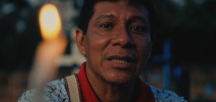 Close de Pepe Manuyama Ahuite, indígena peruano que luta contra a mercantilização da água. Por trás de uma vela, ele olha para a câmera com expressão exasperada. O homem indígena tem cabelos e olhos negros, pele escura e usa uma camisa polo vermelha e azul.