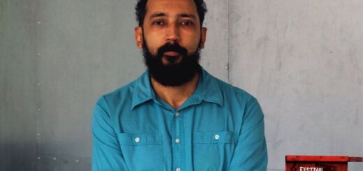 O cineasta Emílio Domingos, pupilo de Patrícia Monte-Mor, olha para a câmera com os braços cruzados sobre o peito. Ele é um homem de meia idade, com barba e cabelos pretos. Ele usa uma camisa de manga comprida azul clara.