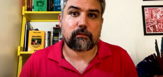 O professor Bruno Malheiro, um homem branco de cabelo e barba grisalhos, olha diretamente para a câmera, com expressão solene, em um frame de um vídeo gravado na sala de sua casa. Ele usa camisa polo vermelha para declarar voto em Lula nas eleições