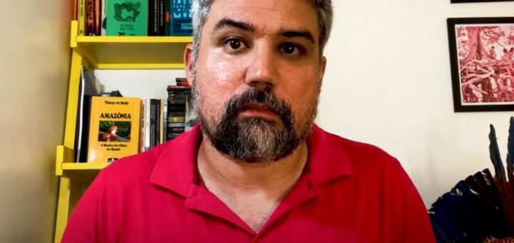 O professor Bruno Malheiro, um homem branco de cabelo e barba grisalhos, olha diretamente para a câmera, com expressão solene, em um frame de um vídeo gravado na sala de sua casa. Ele usa camisa polo vermelha para declarar voto em Lula nas eleições