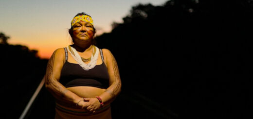 A cacica do povo Akrãtikatêjê, Tônkyre Akrãtikatêjê, em pé sobre os trilhos de uma ferrovia. Tônkyre é iluminada por uma luz que vem de cima, e o sol se põe atrás dela.