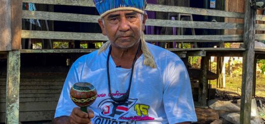 Manoel Munduruku, cacique da aldeia Ipaupixuna, durante o dia, segurando um instrumento tradicional. Ele usa um cocar com penas azuis.