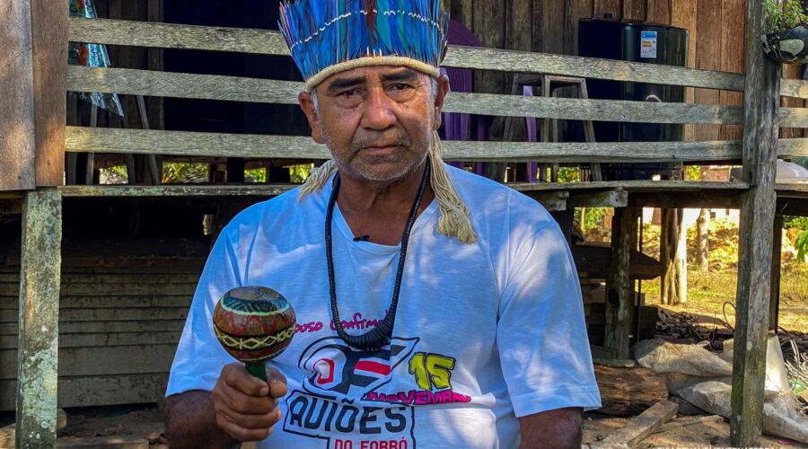 Manoel Munduruku, cacique da aldeia Ipaupixuna, durante o dia, segurando um instrumento tradicional. Ele usa um cocar com penas azuis.
