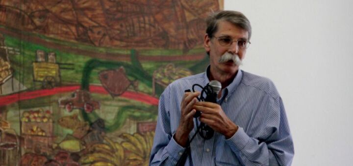 O pesquisador do Inpa Philip Fearnside em frente a uma pintura. Ele usa camisa azul de manga longa e segura um microfone, enquanto olha para o canto direito. Ele tem um proeminente bigode.