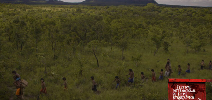 Um grupo de adultos e crianças do povo Xavante caminham na Serra do Roncador. A vegetação na serra é esparsa e algumas montanhas estão no horizonte ao fundo.