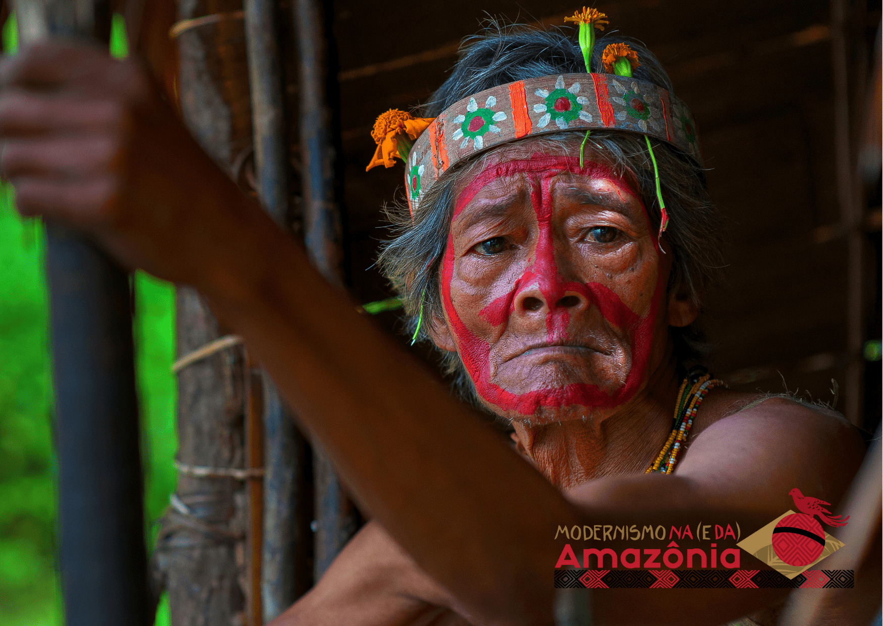 Homem idoso indígena com o rosto pintado de vermelho e uma faixa com imagens de flores ao redor da cabeça.