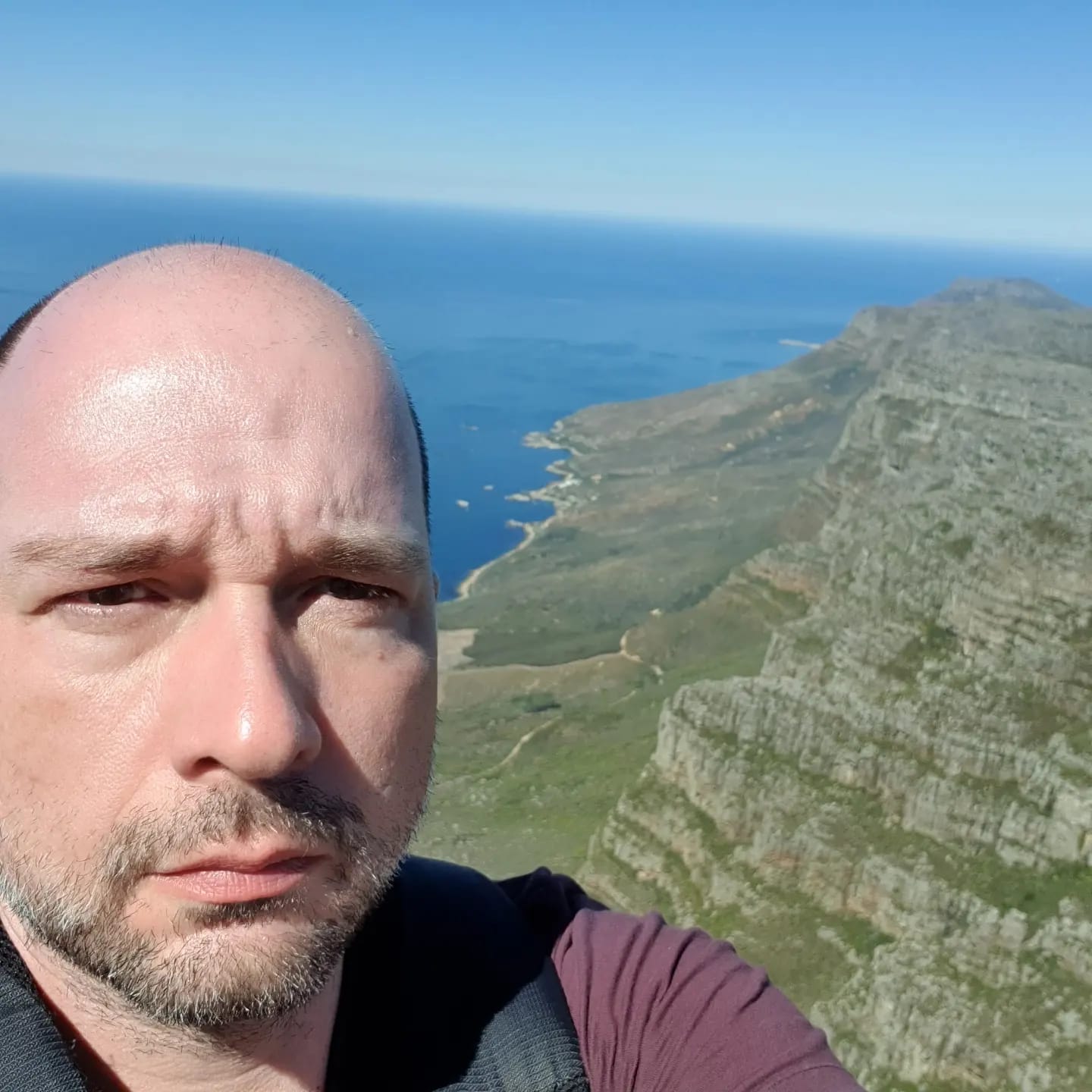 Fabricio Pereira da Silva, professor da Universidade Federal do Estado do Rio de Janeiro, no topo de uma montanha, com a vista de uma praia em um dia ensolarado ao fundo. Fabricio é um homem branco, calvo, e veste uma camiseta roxa.