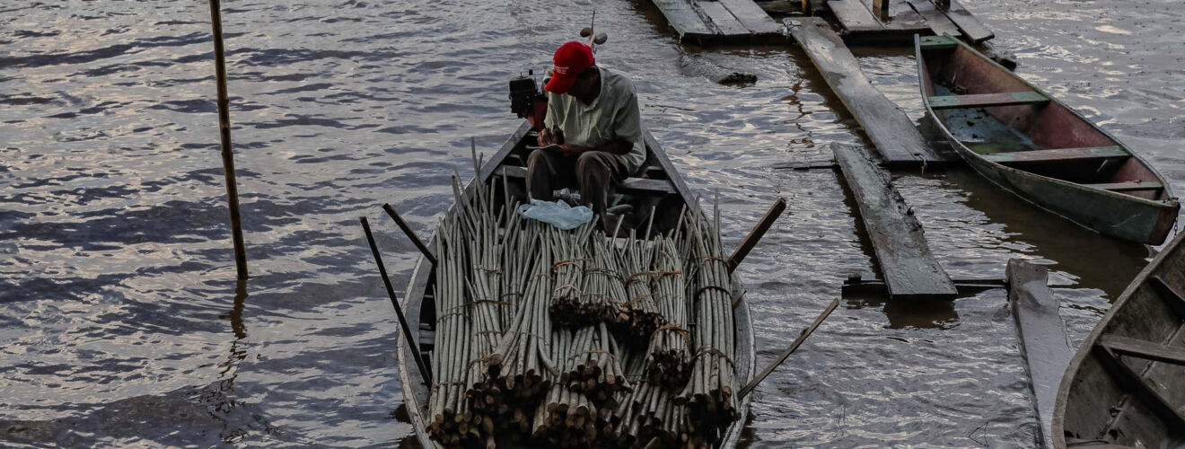 Homem com boné vermelho escrevendo em um caderno. Ele está sentado em uma canoa em um pequeno porto de madeira.
