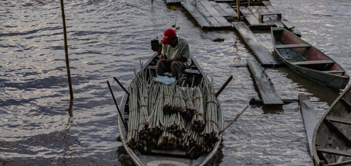 Homem com boné vermelho escrevendo em um caderno. Ele está sentado em uma canoa em um pequeno porto de madeira.
