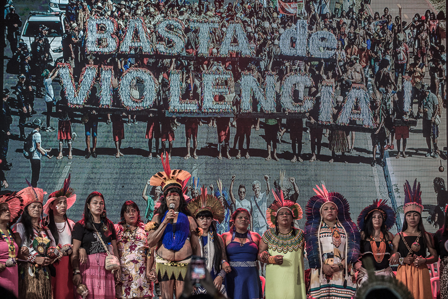 Foto de manifestação indígena no Acampamento Terra Livre 2022. Em fileira, mulheres com cocares falam ao microfone sob um cartaz que se lê "Basta de Violência".