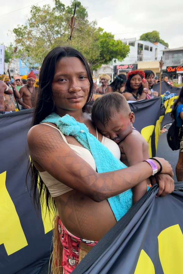 Em um retrato que simboliza a luta pela vida, esta mãe indígena carrega seu filho sob o sol de Belém, levando a força de seu povo e a esperança de um futuro seguro. Foto: Alice Palmeira.