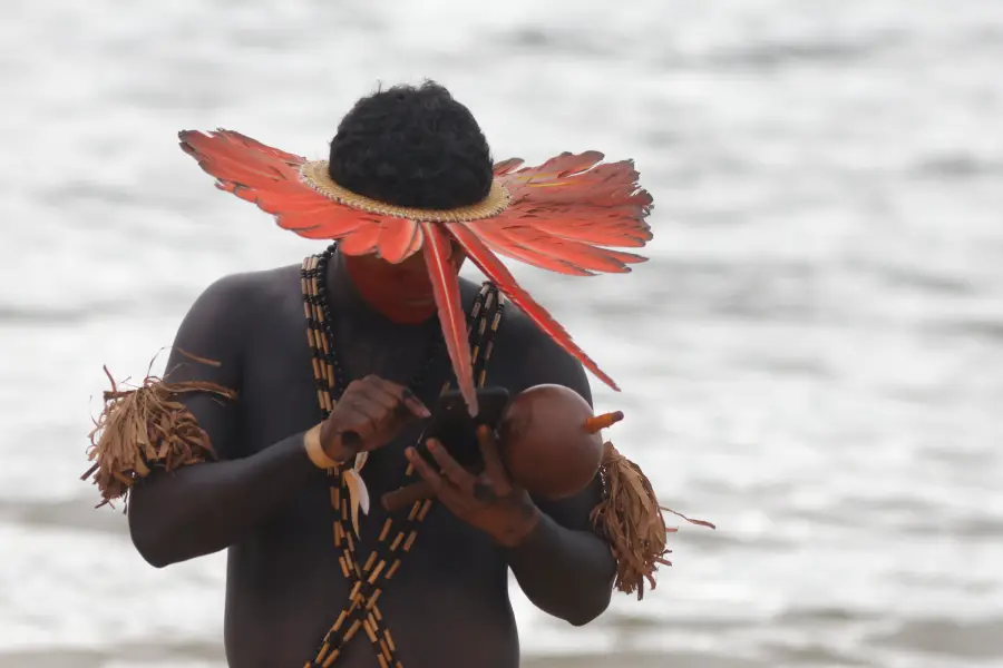 Indígena de cocar e maracá consulta o celular nas margens do Xingu, retratando a contemporaneidade das comunidades originárias. Foto: Oswaldo Forte / Amazônia Latitude.