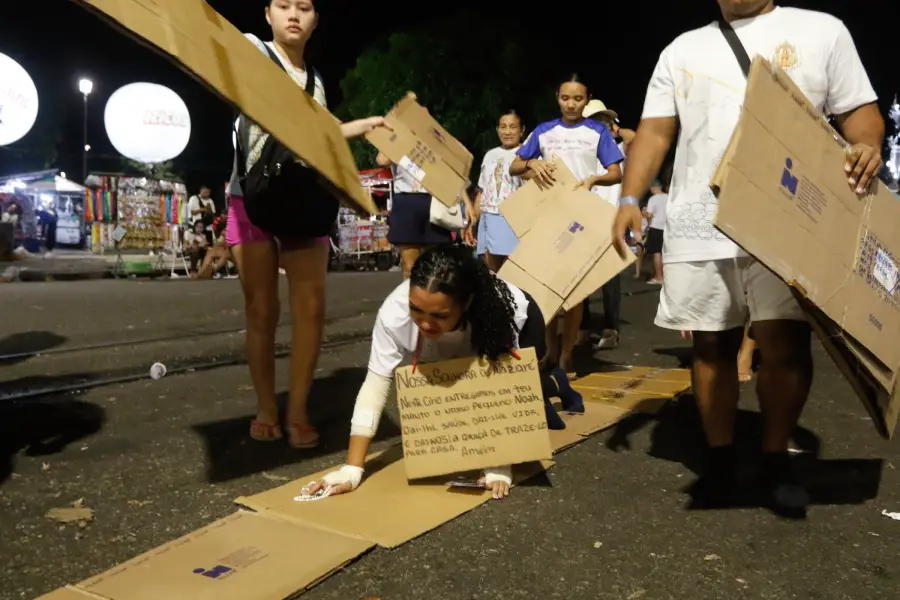 Em um ato de sacrifício e devoção, uma romeira, com os joelhos e mãos protegidos por curativos, rasteja sobre papelões no asfalto. Em suas mãos, um terço e uma placa com um pedido comovente a Nossa Senhora de Nazaré pelo filho. Foto: Oswaldo Forte/Amazônia Latitude.
